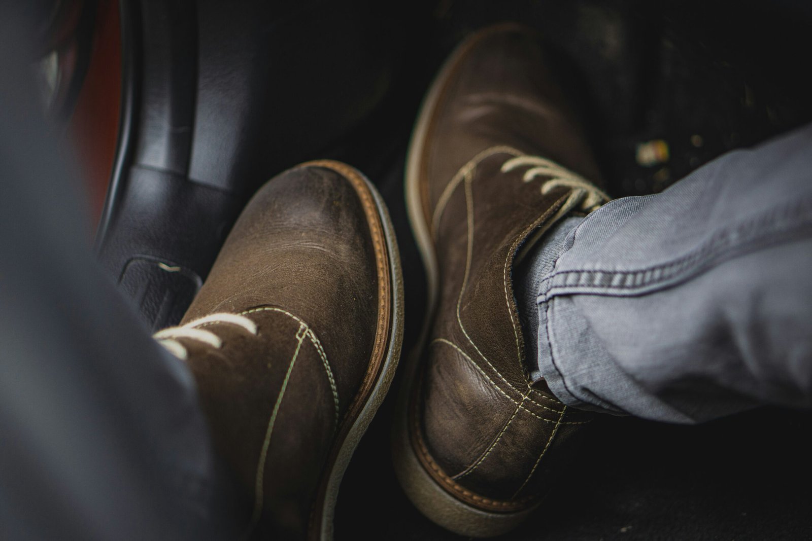 Brown leather casual shoes on a person's feet while sitting in a car, shown close-up with soft lighting and visible wear on the leather.